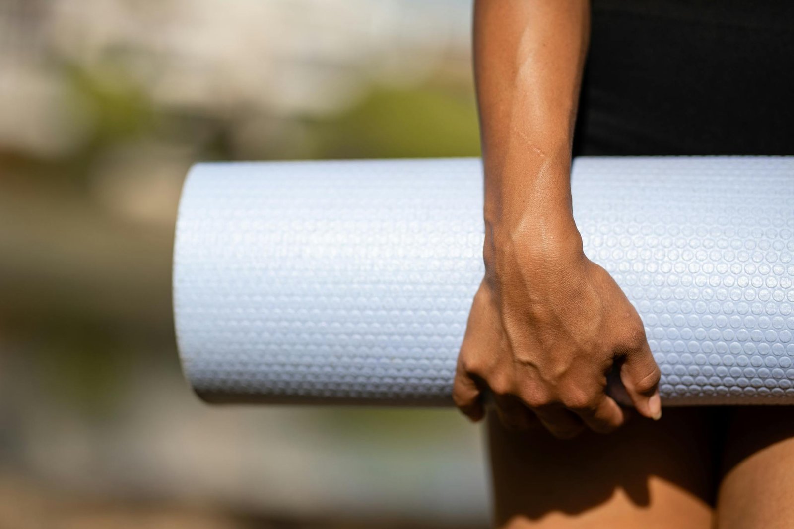A person holding a rolled white yoga mat outdoors with a blurred background, signifying fitness and exercise.